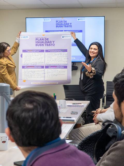 En una reunión, dos mujeres presentan un plan en un póster mientras los demás observan atentamente.