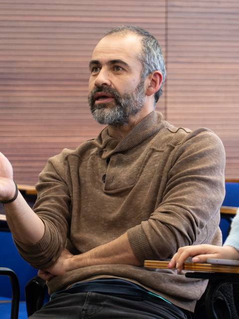Un hombre con barba y suéter conversando en un aula.