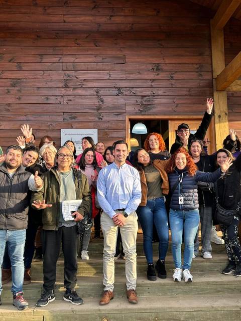Un grupo de personas sonrientes y levantando las manos frente a una casa de madera.