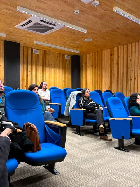 Un grupo de personas está sentado en una sala con sillas azules, prestando atención a una presentación.