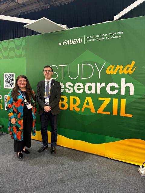 Dos personas posando frente a un gran cartel que promociona estudios e investigación en Brasil.