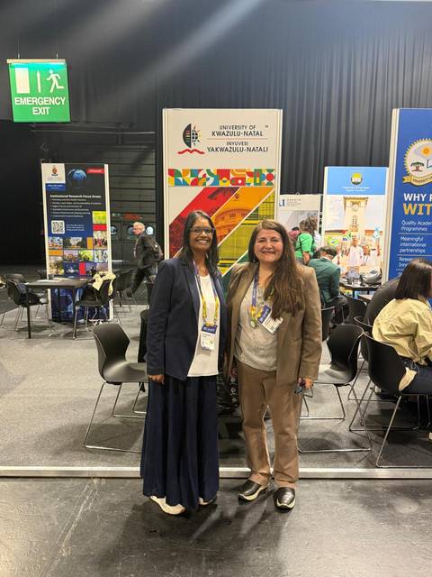 Dos mujeres posando sonrientes en un evento con stands variados detrás.