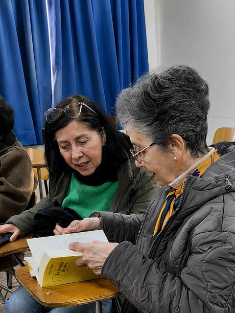Dos mujeres conversan animadamente mientras revisan un libro en un aula.