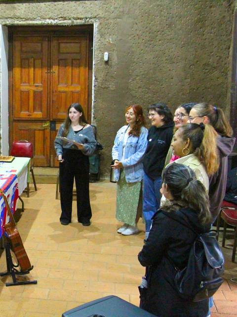 Un grupo de personas escucha a una mujer que habla frente a una mesa decorada con comida típica chilena.