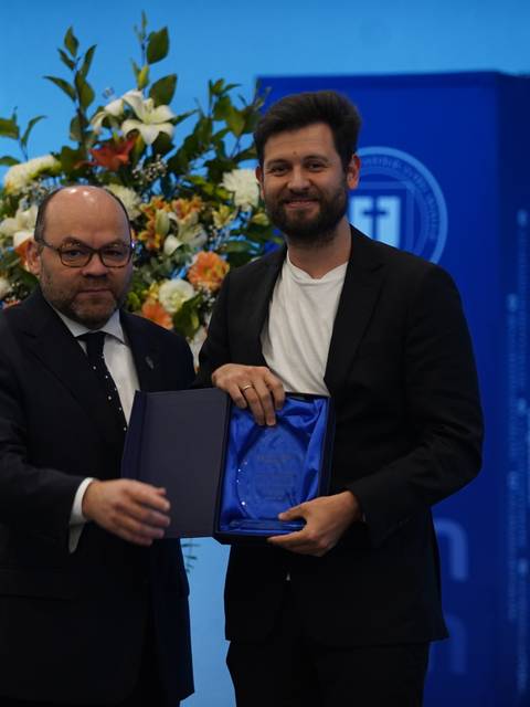 Dos hombres posando en un evento con un fondo decorado y un premio en sus manos.
