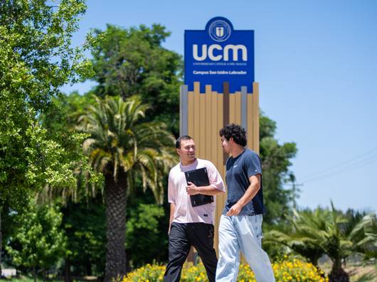 Dos jóvenes conversan alegremente frente a la entrada del campus de la UCM en un día soleado.