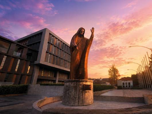 Una impresionante escultura de un figura religiosa se alza frente a un edificio moderno bajo un cielo colorido al atardecer.