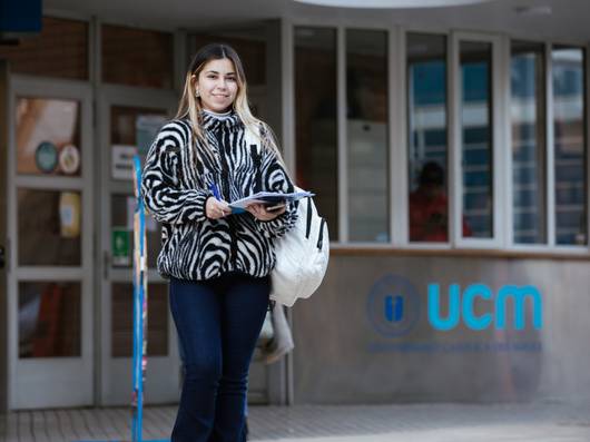 Una estudiante sonriente con un abrigo de cebra que sostiene unos papeles frente a la Universidad Católica del Maule.