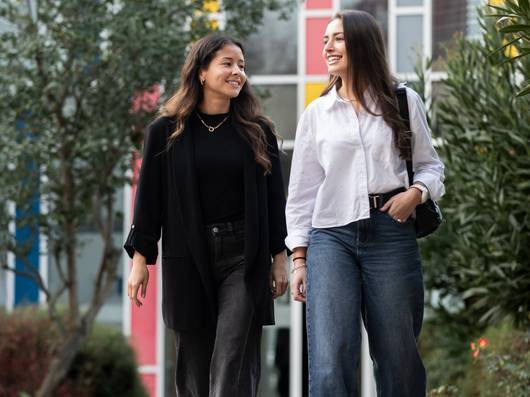 Dos amigas caminan juntas sonrientes por un camino rodeado de vegetación y coloridos muros en un entorno urbano.