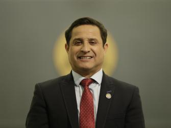 Un hombre sonriente con un traje formal y una corbata roja, de fondo difuso.