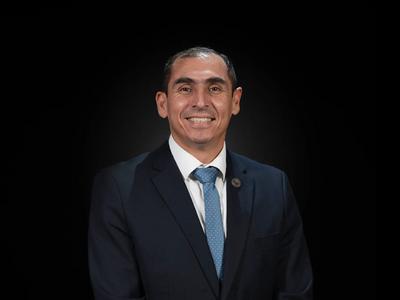 Un hombre sonriente vestido con un traje oscuro y corbata en un fondo negro.