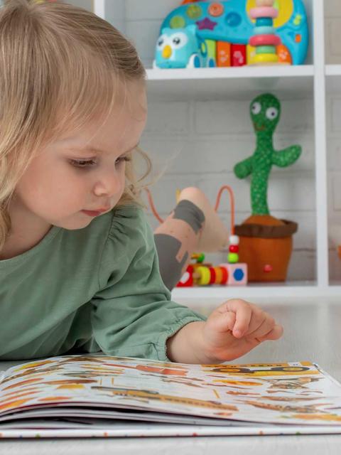 Niña pequeña leyendo un libro en el suelo rodeada de juguetes.
