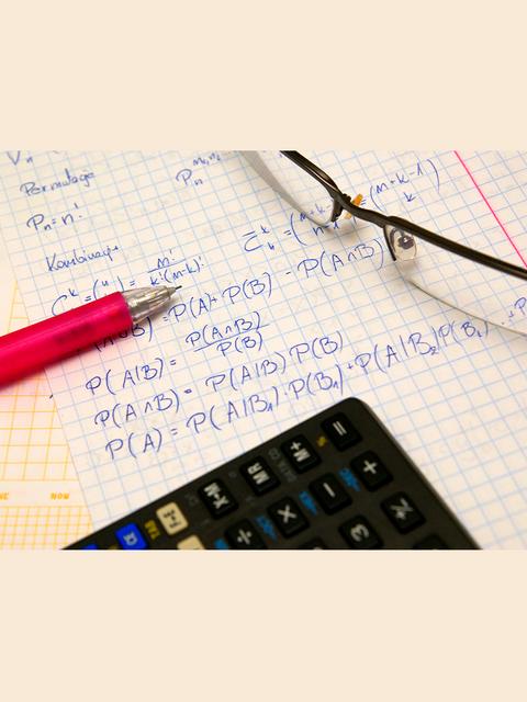 En la imagen se ven fórmulas matemáticas escritas en una hoja de cuaderno, junto a un calculador y un lápiz rojo.