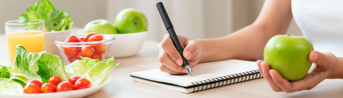 Una persona escribe en un cuaderno mientras sostiene una manzana verde, rodeada de ensalada, tomates y un vaso de jugo de naranja.