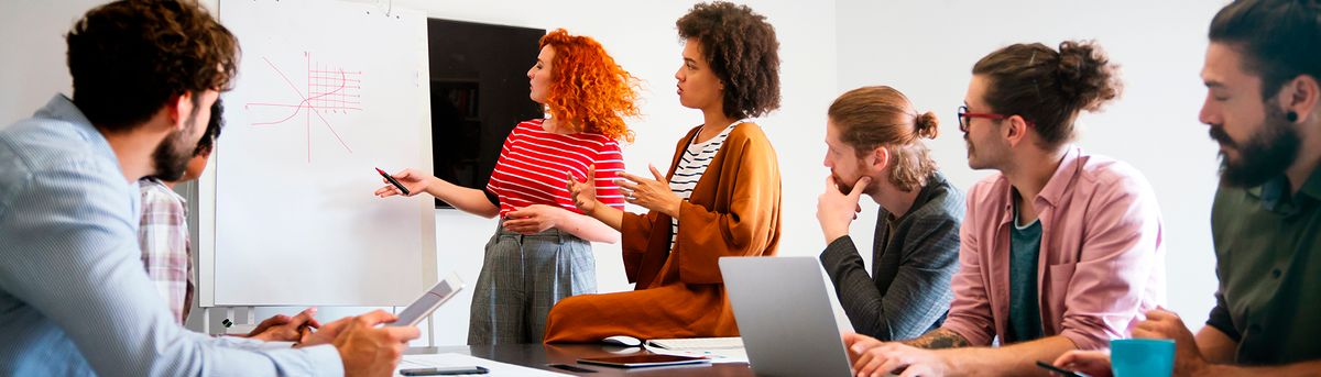 Un grupo de personas participa en una reunión en una oficina, mientras una mujer presenta información en una pizarra.