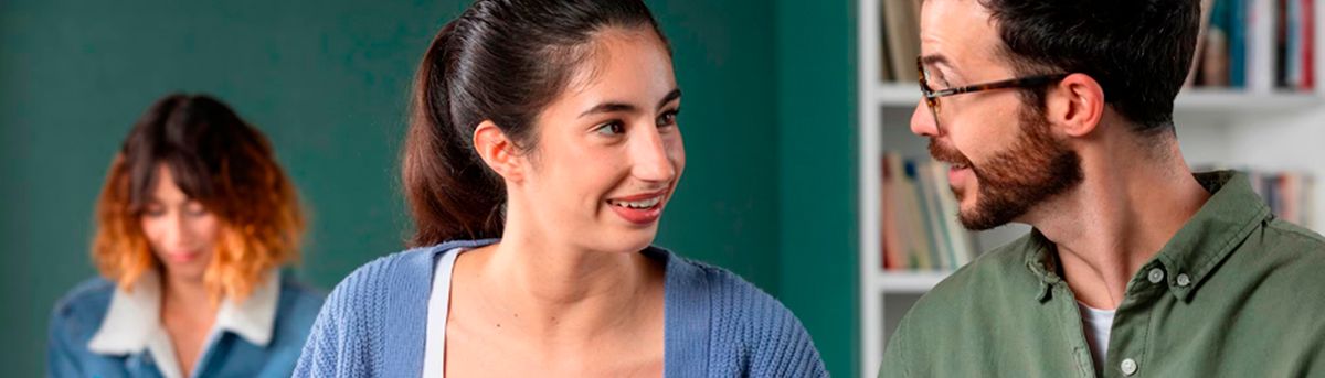 Una mujer y un hombre conversan mientras una mujer con cabello rizado se encuentra al fondo.