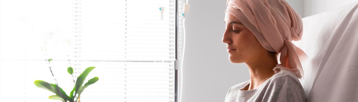 Una mujer con un pañuelo rosa en la cabeza, sentada en una cama de hospital, mirando por la ventana.