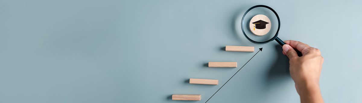 Una mano sostiene una lupa enfocando un símbolo de graduación sobre unas escaleras de madera que representan el progreso académico.