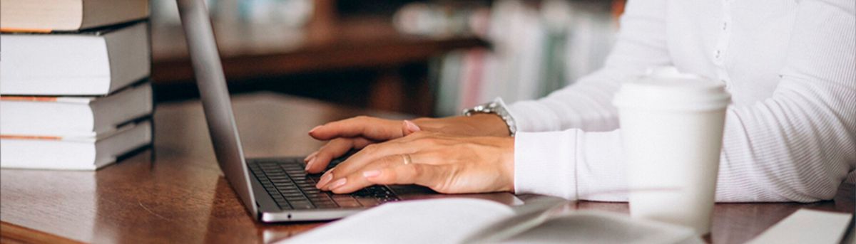 Una persona teclea en un ordenador portátil mientras tiene una taza de café a un lado y libros apilados.