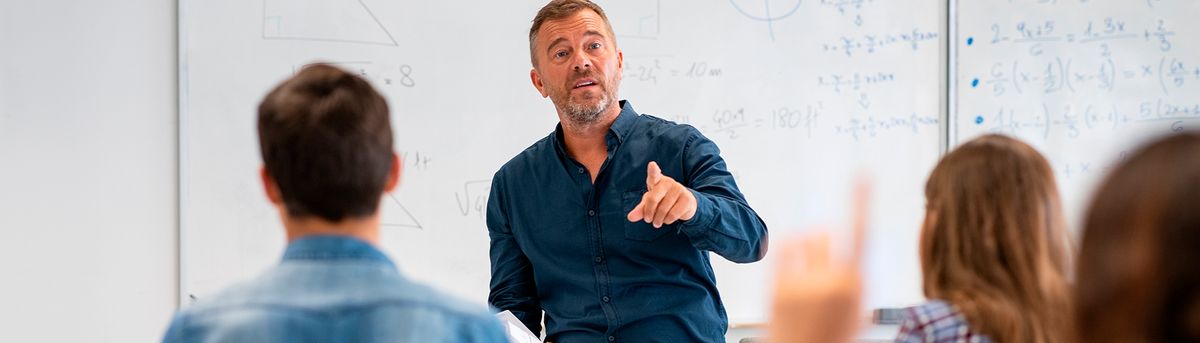 Un profesor está explicando un tema frente a su clase, mientras un estudiante levanta la mano para participar.
