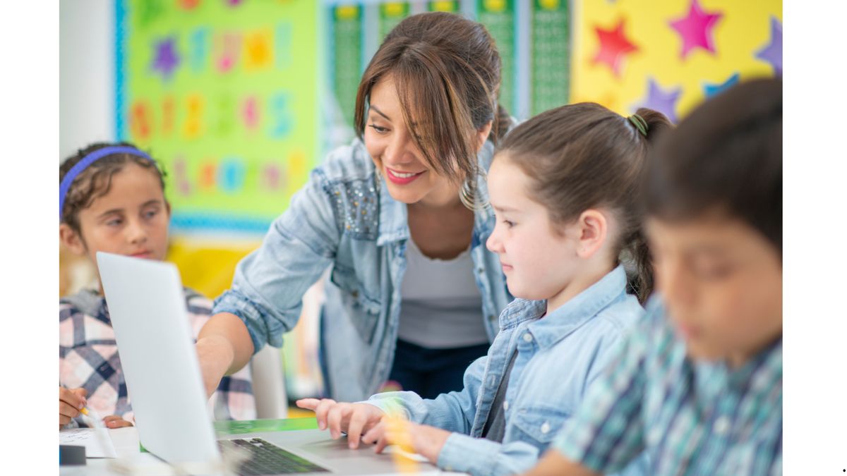 Una profesora ayuda a dos niñas con la computadora en una sala de clases colorida.