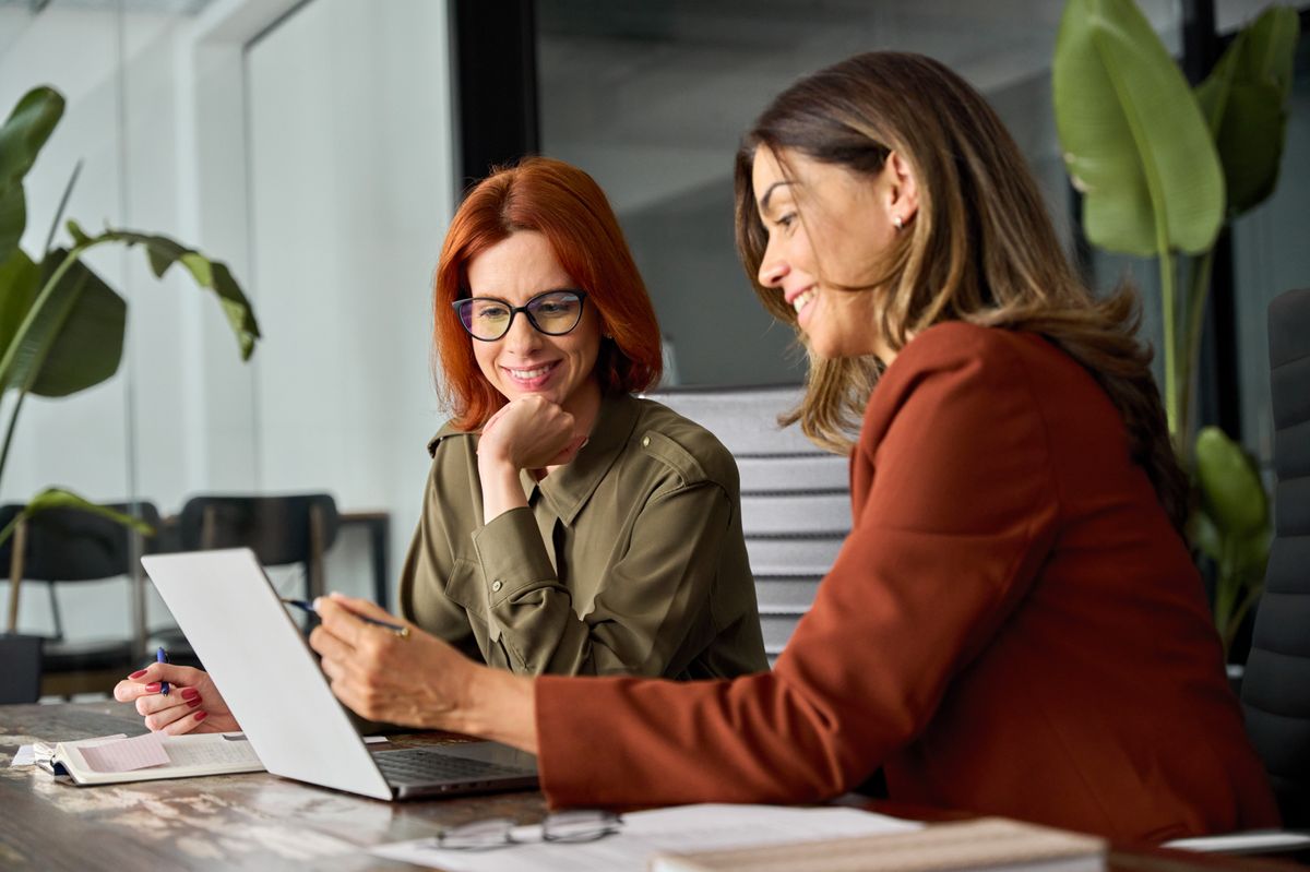 Dos mujeres se encuentran conversando animadamente frente a una laptop en una oficina moderna.