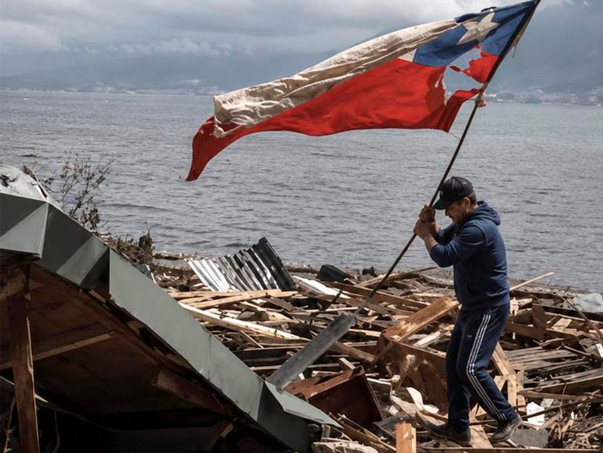 Un hombre sostiene con fuerza la bandera chilena frente a escombros tras un desastre natural.