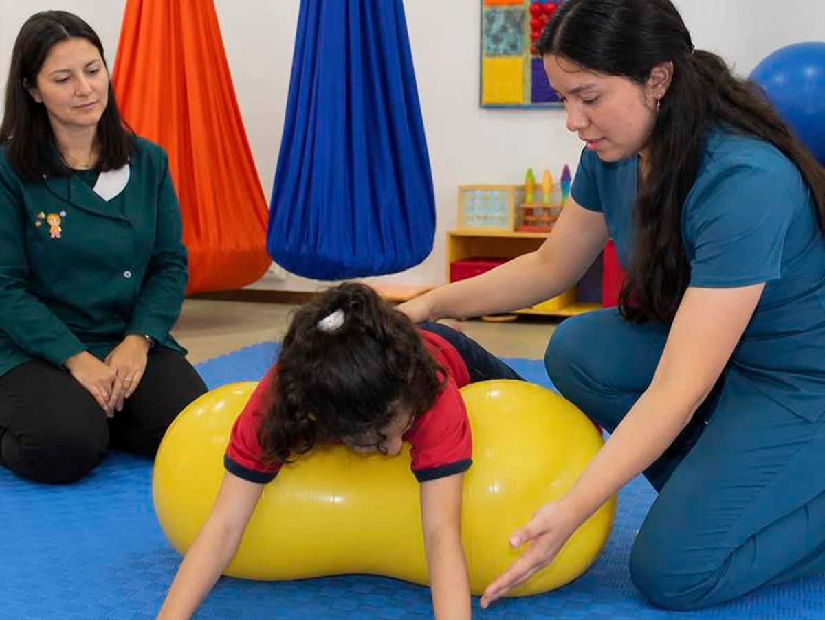 Una terapeuta ayuda a un niño a jugar sobre una pelota mientras otra mujer observa en un ambiente de terapia.