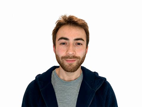 Un joven sonriente con barba, vestido con una polera y un chaleco.