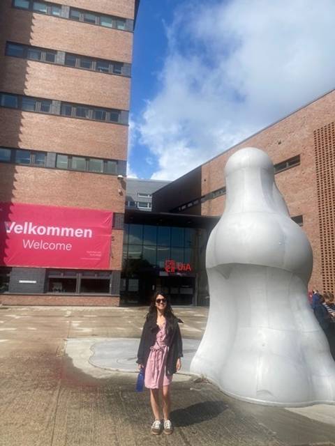 Una persona posando frente a un edificio moderno con una escultura abstracta y un cartel de bienvenida.