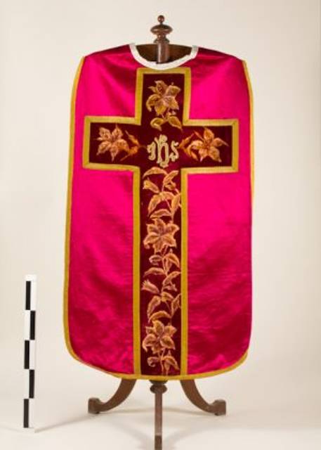 La imagen muestra un alba de color fucsia con un diseño de cruz y bordados florales en tonos dorados.