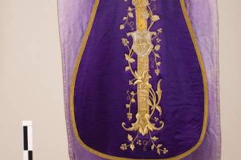 La imagen muestra un objeto textil de color púrpura con bordados dorados y detalles ornamentales.