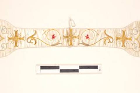 Se trata de una banda litúrgica decorada con hilos dorados y bordados rojos sobre un fondo claro.