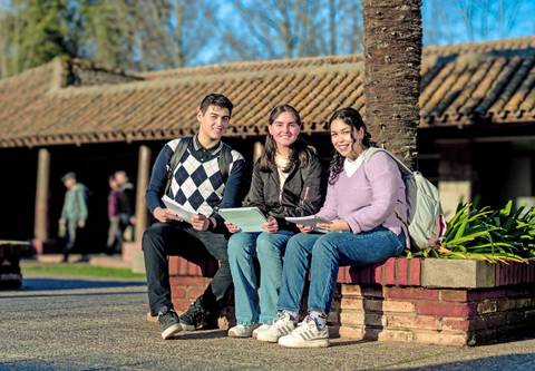 Tres estudiantes se sientan en un banco al aire libre, sonriendo y conversando mientras sostienen tabletas y papeles.