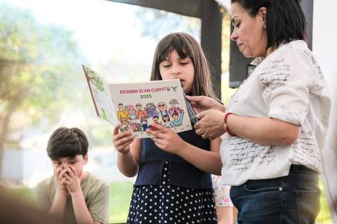 Una niña lee su cuaderno de escritura mientras una adulta la acompaña.