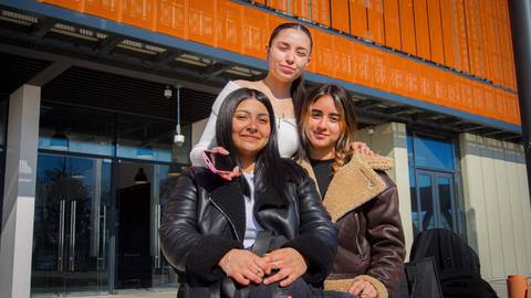 Tres amigas posando juntas con sonrisas en un día soleado frente a un edificio moderno.