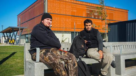 Dos amigos sentados en un banco frente a un edificio moderno en un día soleado.