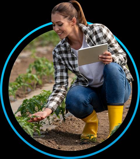Una mujer sonriente se agacha cuidando plantas mientras sostiene una tablet.