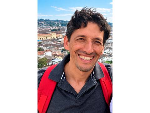 Un hombre sonriente con una polera negra y una mochila roja mirando hacia la cámara, con una hermosa vista de la ciudad de fondo.