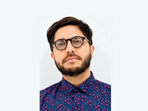 Un hombre con barba y gafas, viste una camisa con flores en un fondo claro.