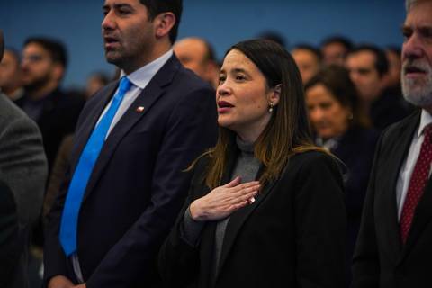 Un grupo de personas en un acto cívico, donde una mujer lleva la mano en el corazón durante el himno nacional.