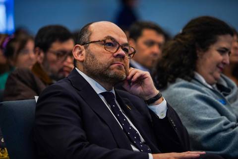 Un hombre con anteojos escucha atentamente en un evento con público detrás.