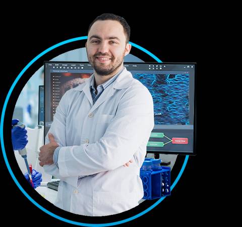 Un hombre sonriente en un laboratorio, vestido con una bata blanca y de pie frente a una computadora mostrando gráficos.