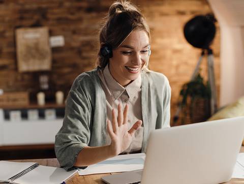 Una mujer sonriente, con audífonos, saludando a través de la computadora mientras trabaja desde casa.