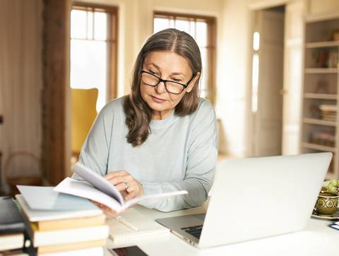 Una mujer lee un libro sentada en una mesa con una computadora portátil y varios libros a su alrededor.