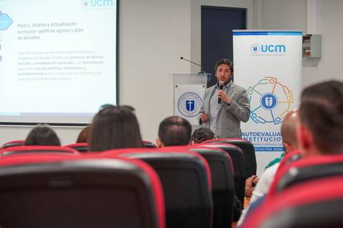 Un expositor presenta un tema académico frente a un grupo de personas en un salón de clases.