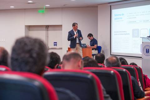 Un presentador expone sobre un sistema de gobierno frente a una audiencia en un auditorio.