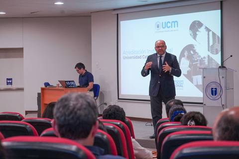Un expositor da una charla sobre acreditación en una universidad mientras el público escucha atentamente.