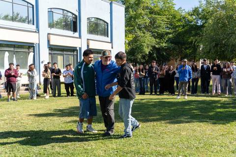 Tres personas se están saludando en un evento al aire libre mientras un grupo de personas los observa.