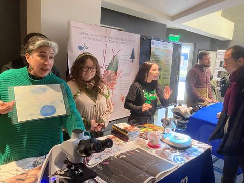En una feria científica, personas presentan proyectos y materiales sobre biología, rodeados de microscopios y gráficos.